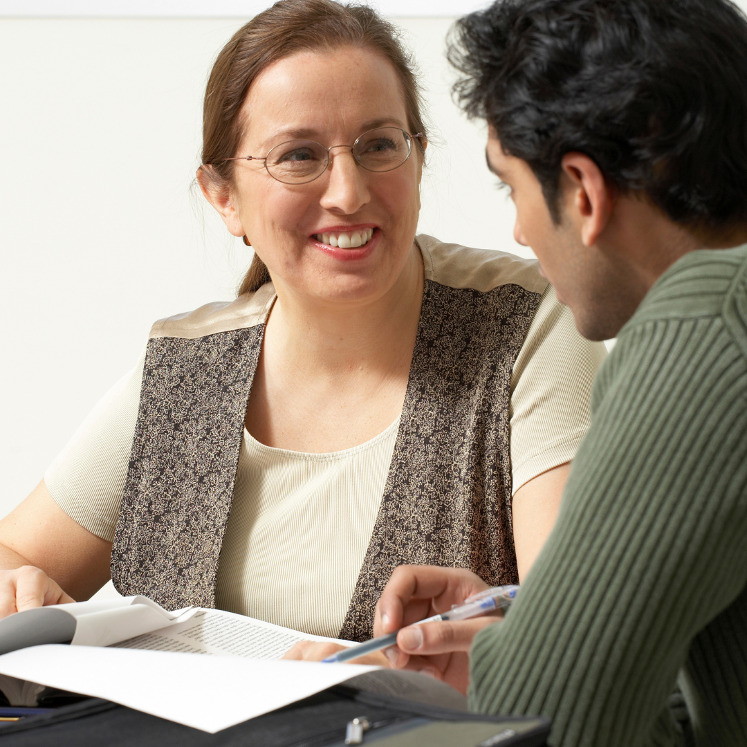 Image of White female instructor sitting next to a male student helping the student with a question. The instructor and student are making eye contact and the instructor has a supportive, smiling appearance.